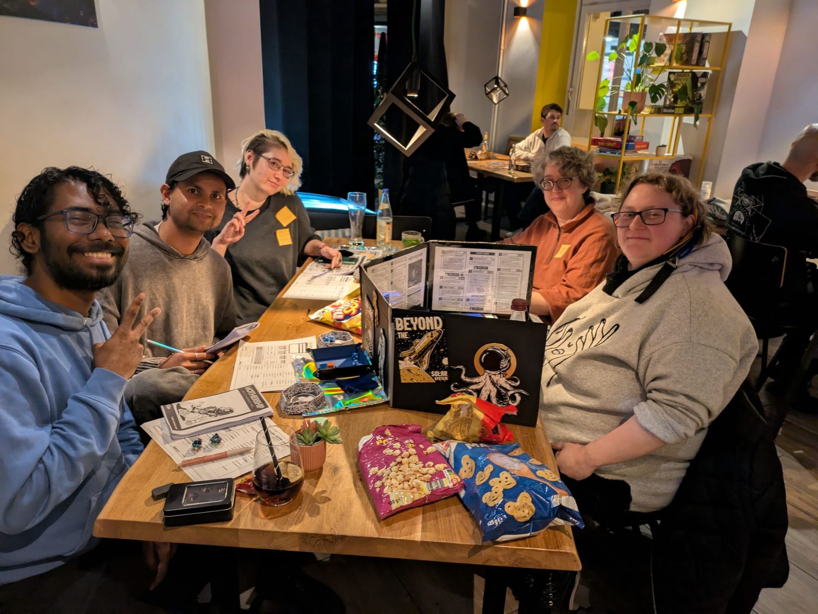 Five players either side of a hand-decorated game master screen, crisp packets open on the table.