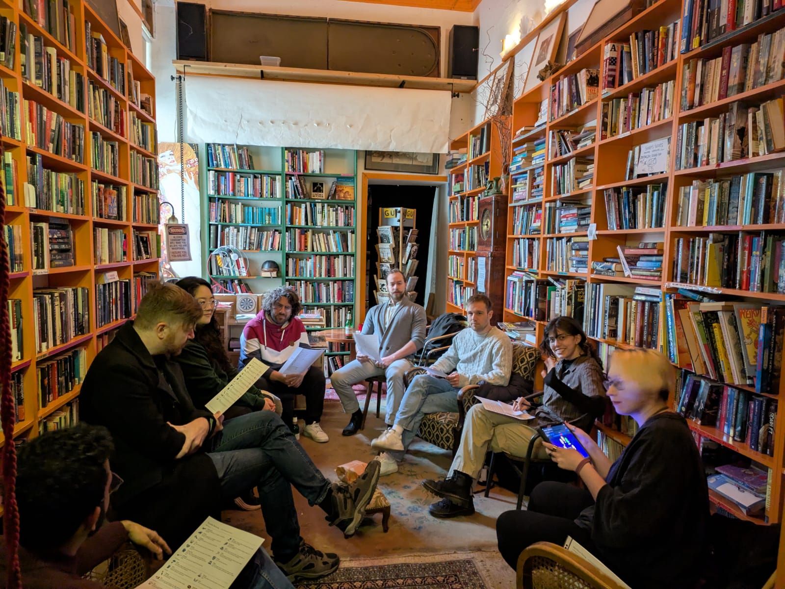 Ten people sitting in a circle of armchairs between tall bookshelves, character sheets in their laps.