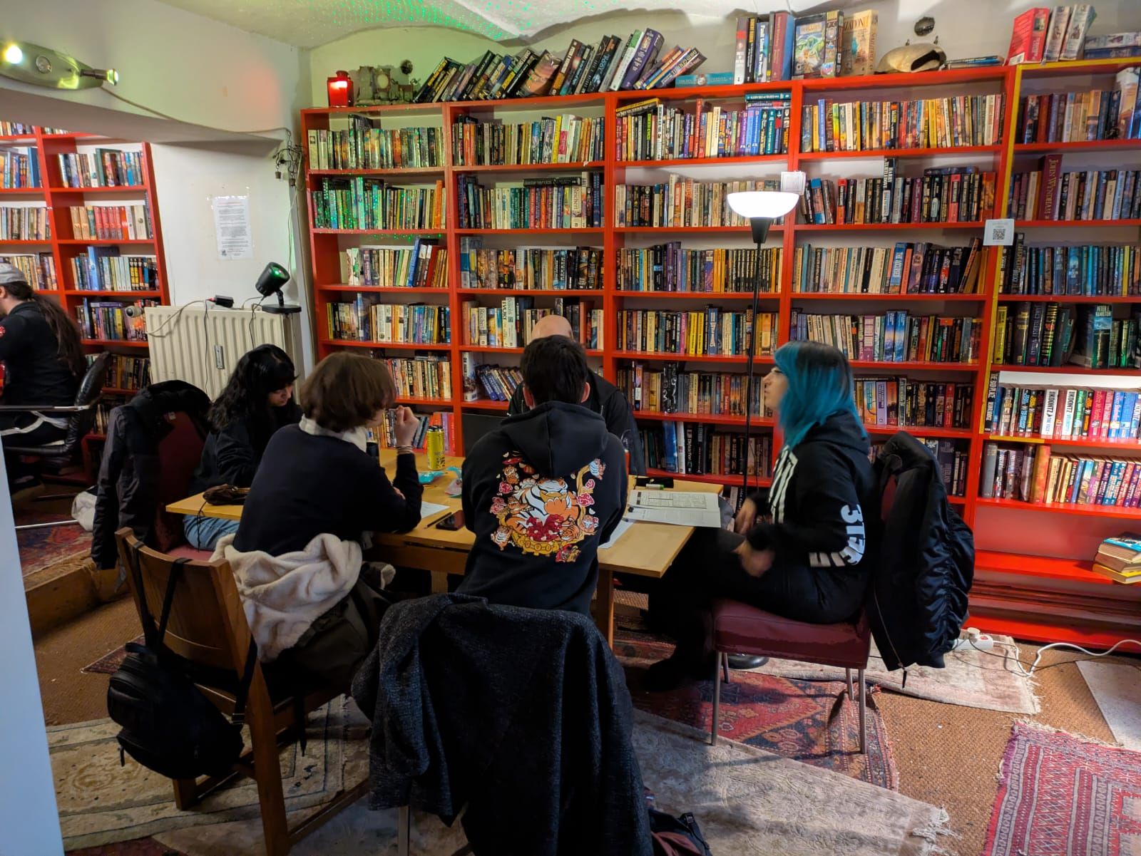 Four players at a long table in front of floor-to-ceiling red bookshelves, one leaning over a character sheet.