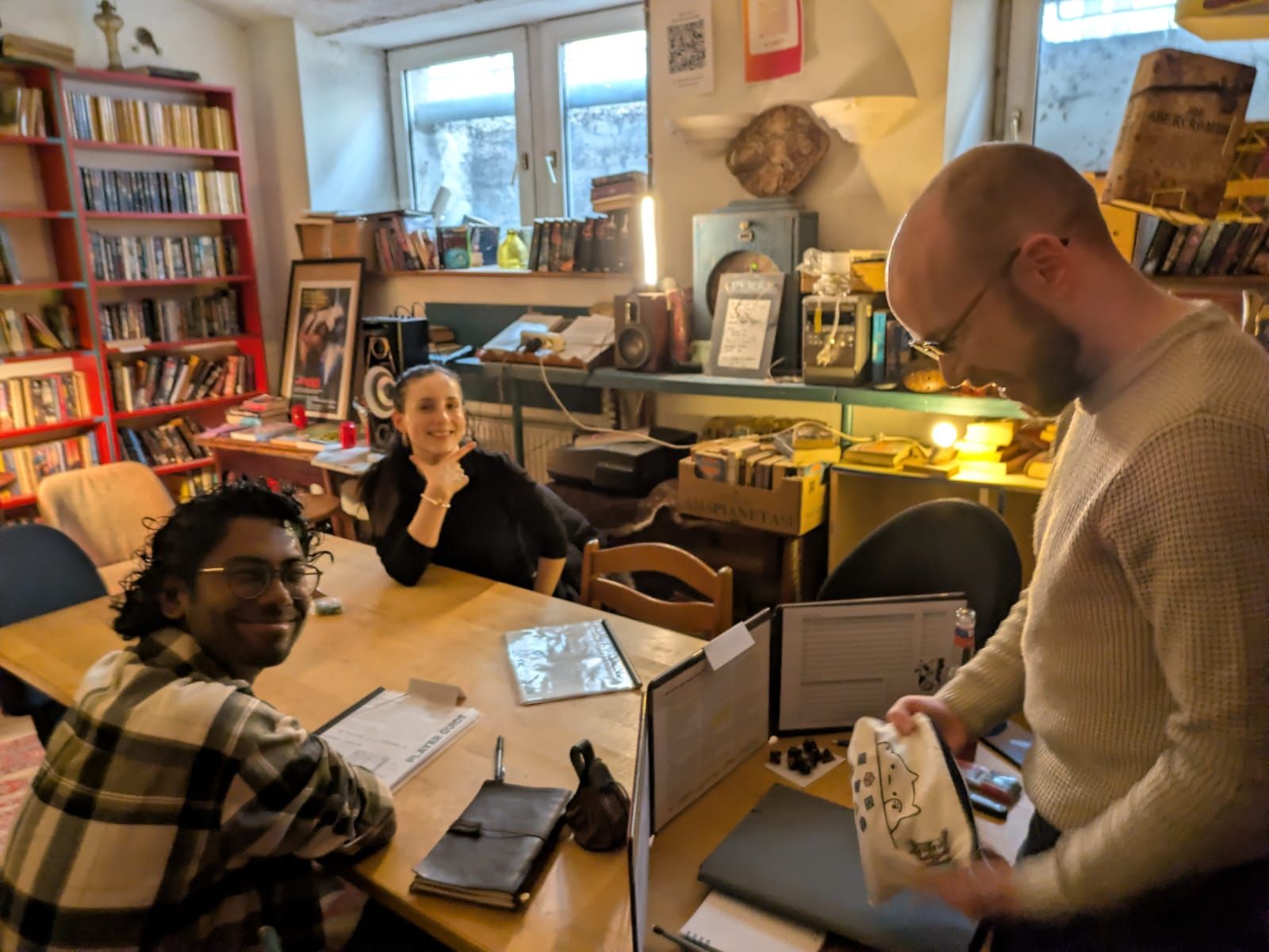A game master standing at the head of a table opening a cloth dice bag, two players looking up and smiling.