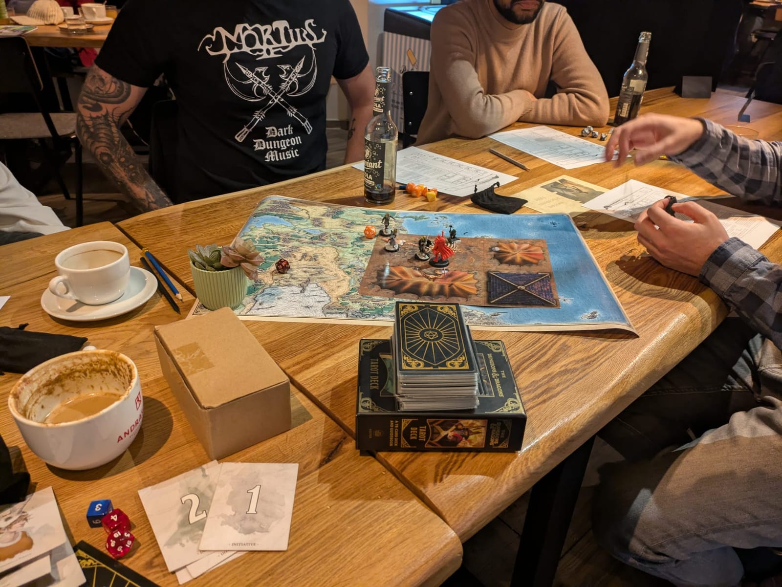 Close-up of a table mid-game: a hand-drawn map with miniatures, a tarot-style card deck, dice, table numbers and a coffee cup.