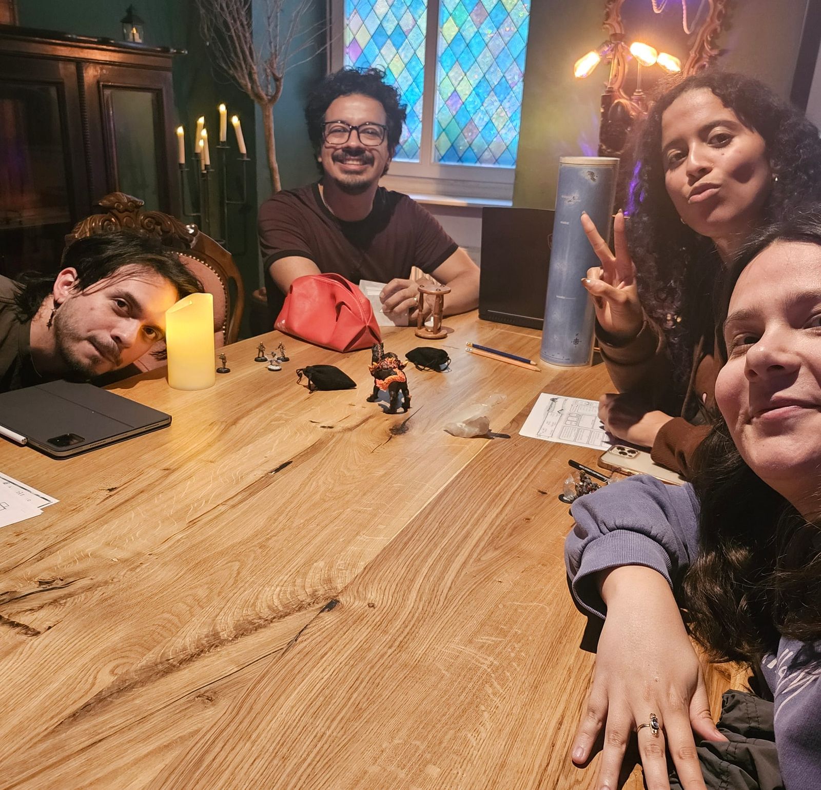Miniatures and dice bags on a wooden table by a stained glass window, four players leaning into the camera.
