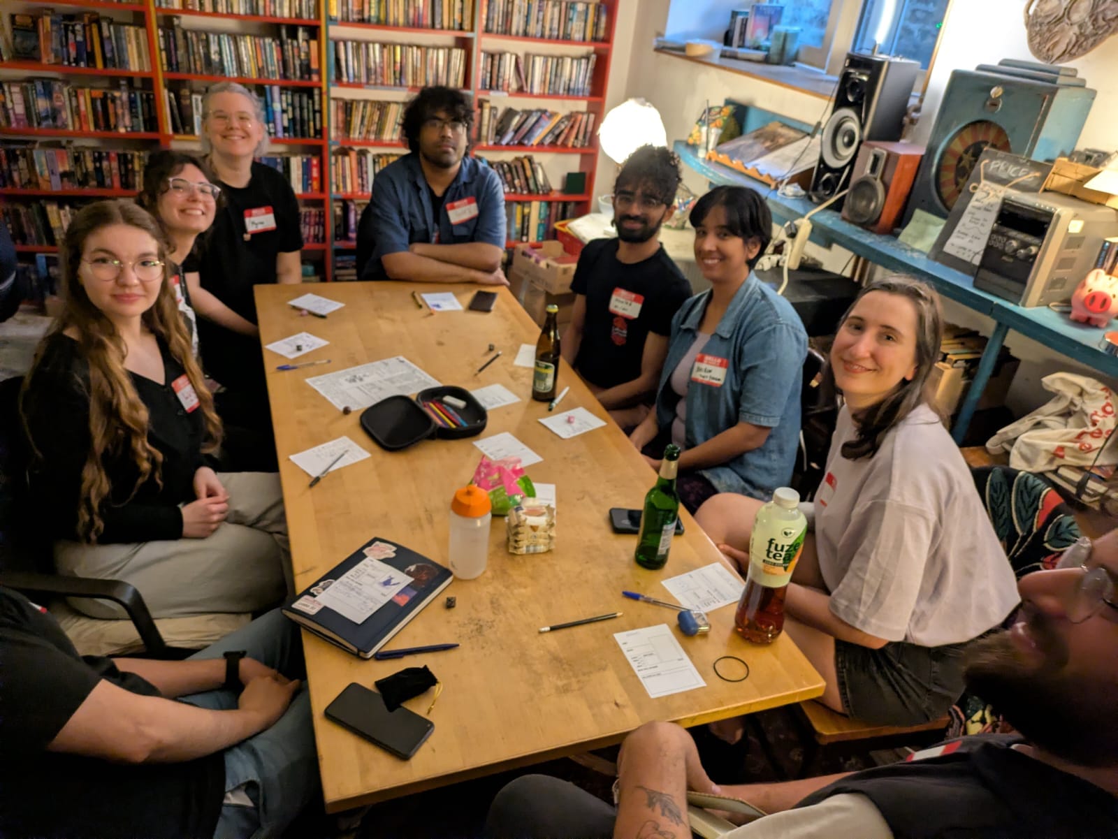 Seven players wearing name stickers around a wooden table, smiling toward the camera, papers and drinks in front of them.