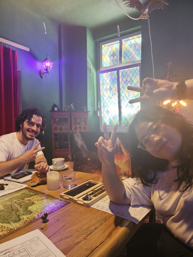 A player grins and makes a peace sign across a wooden table with a map, under a stained-glass window and a purple lamp.