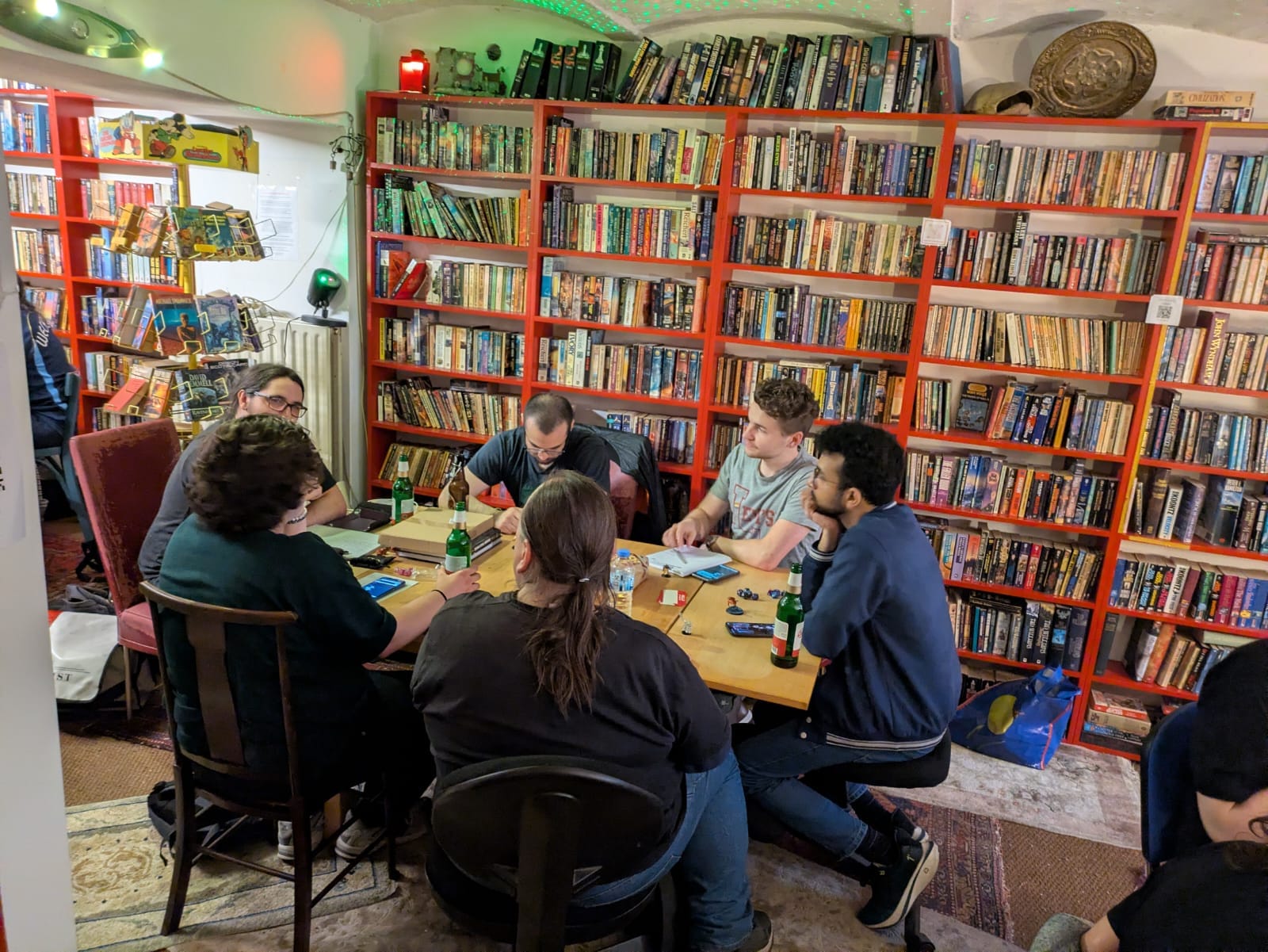 Players leaning over a small table in front of floor-to-ceiling red bookshelves, one pointing at the table.