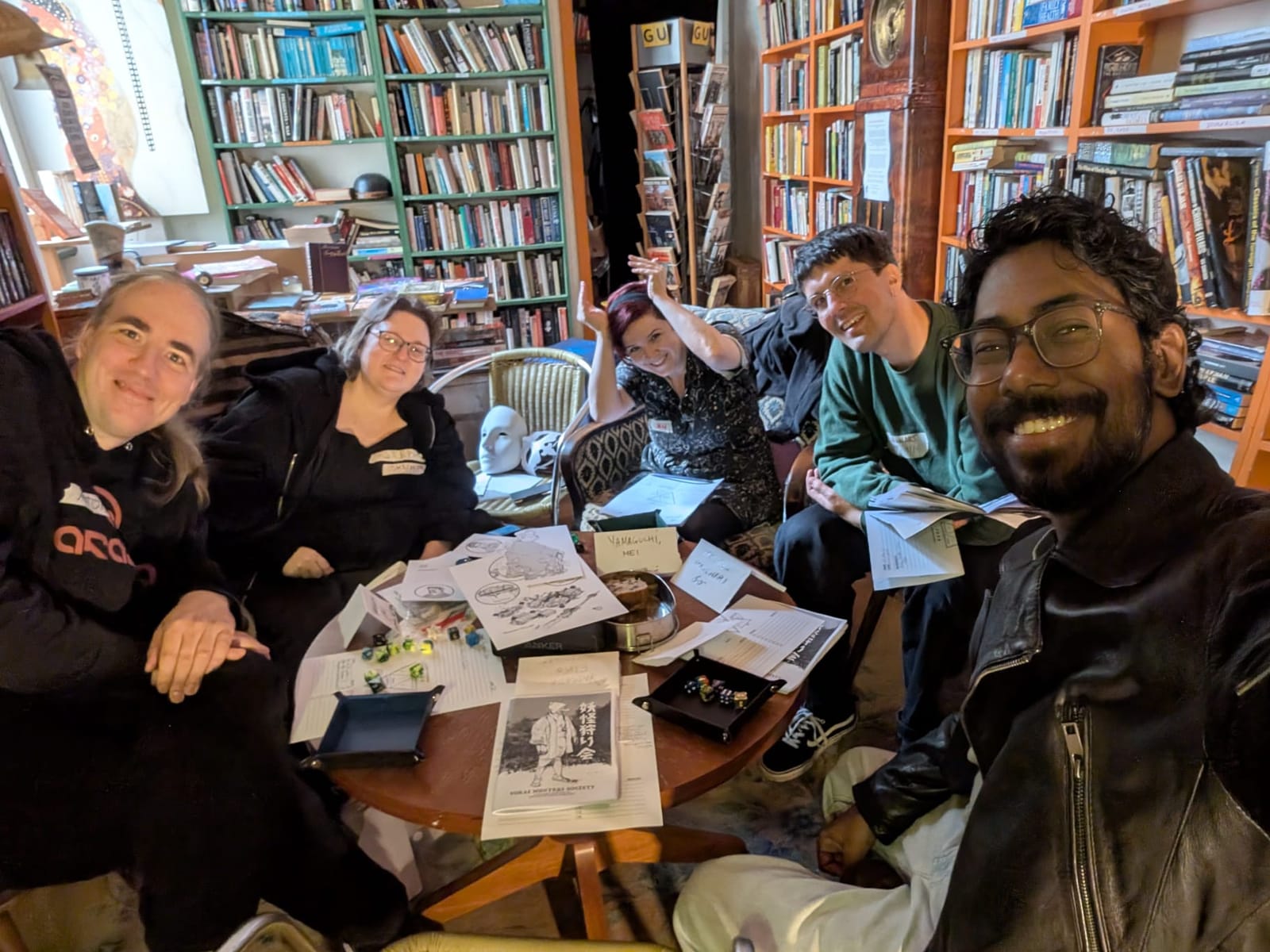 A selfie of five smiling players around a small round table covered in papers and dice, bookshelves behind them.