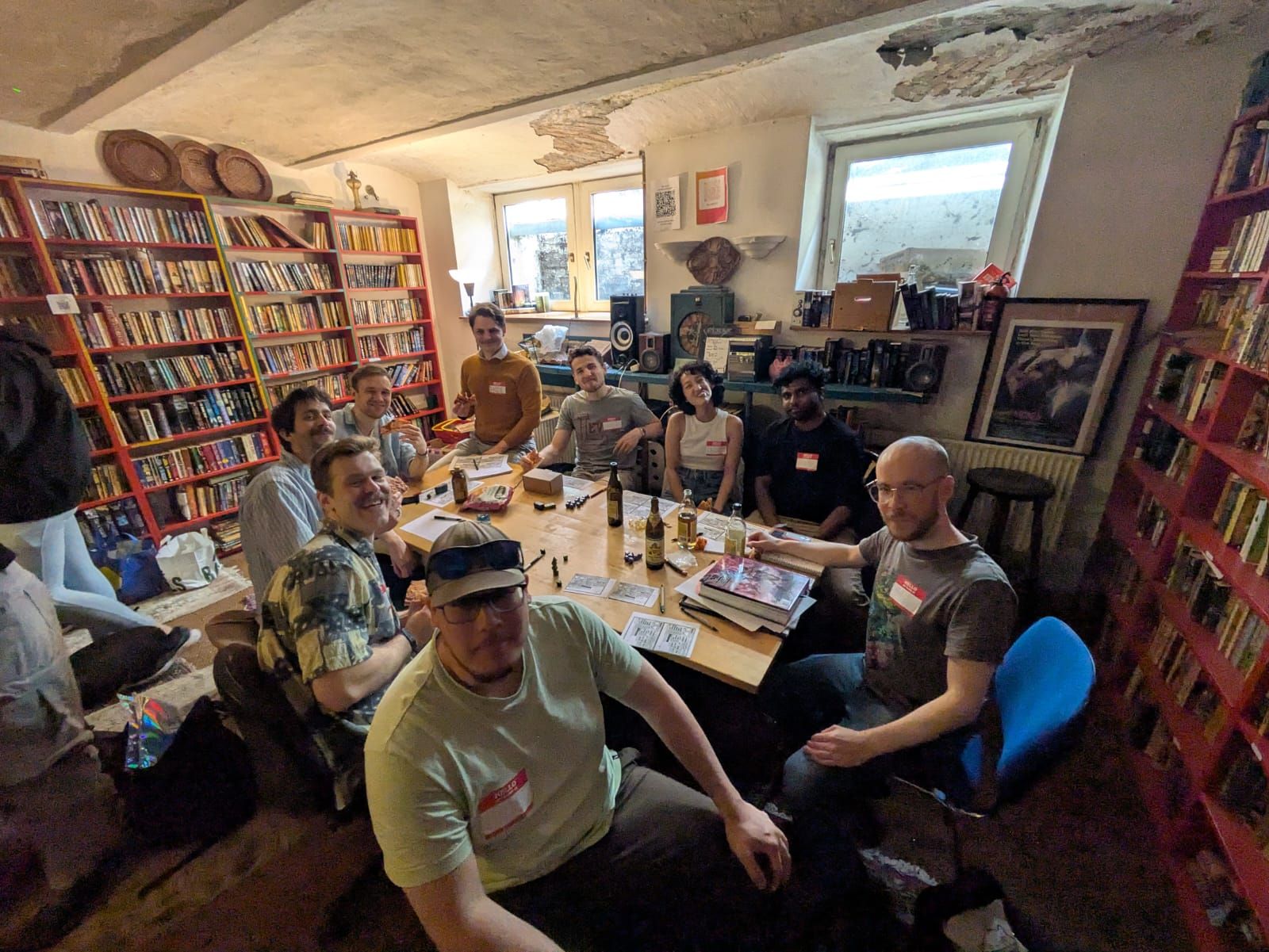 Ten people around one long table in a book-lined basement, name badges on, bottles and food between the dice.