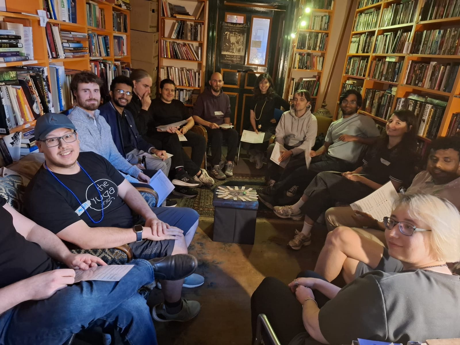 A crowded circle of people on sofas and armchairs between bookshelves, papers in their laps, seen from the front row.
