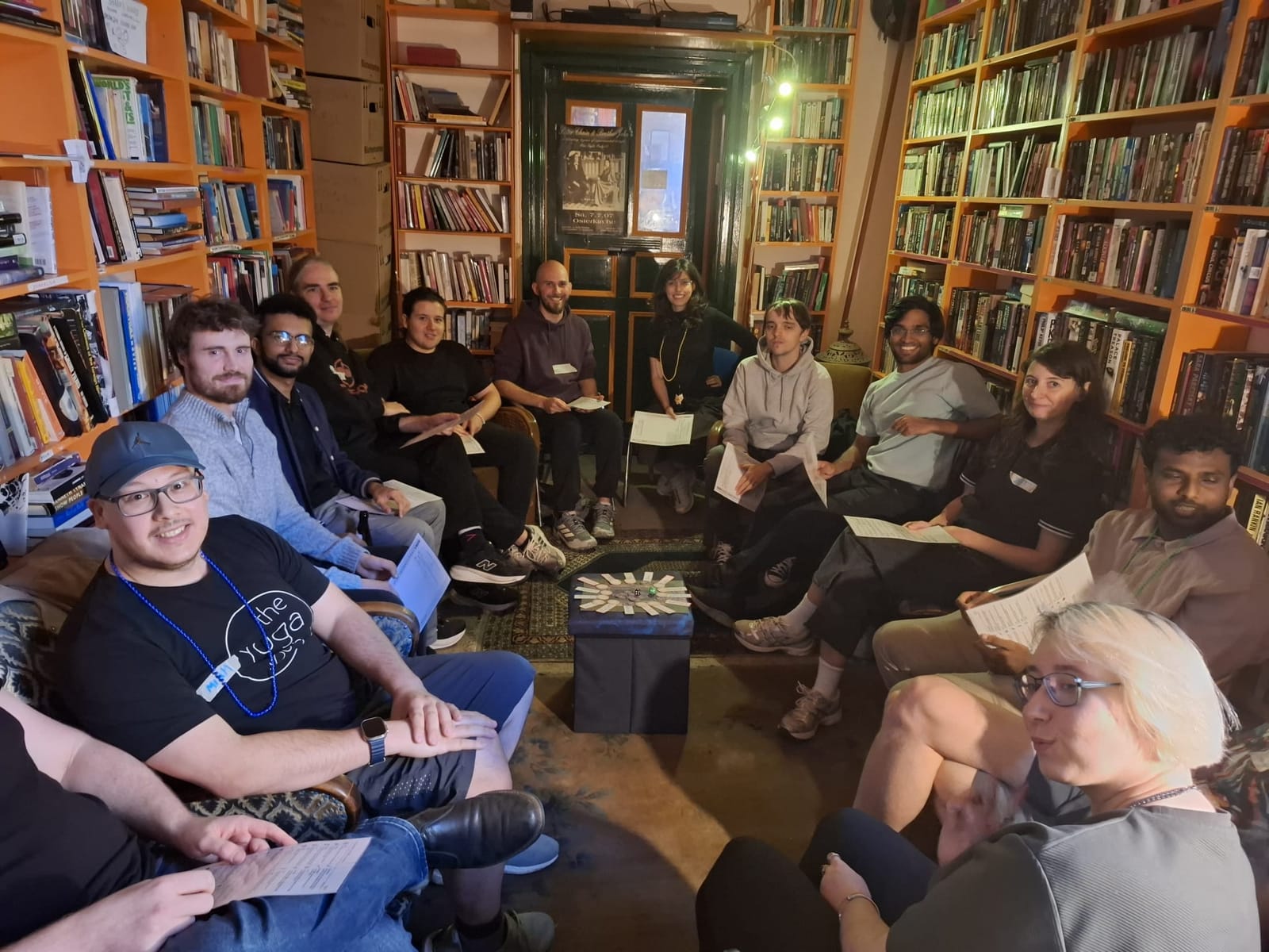 A wide circle of about fifteen people seated between tall bookshelves, holding printed sheets, a footstool in the middle.