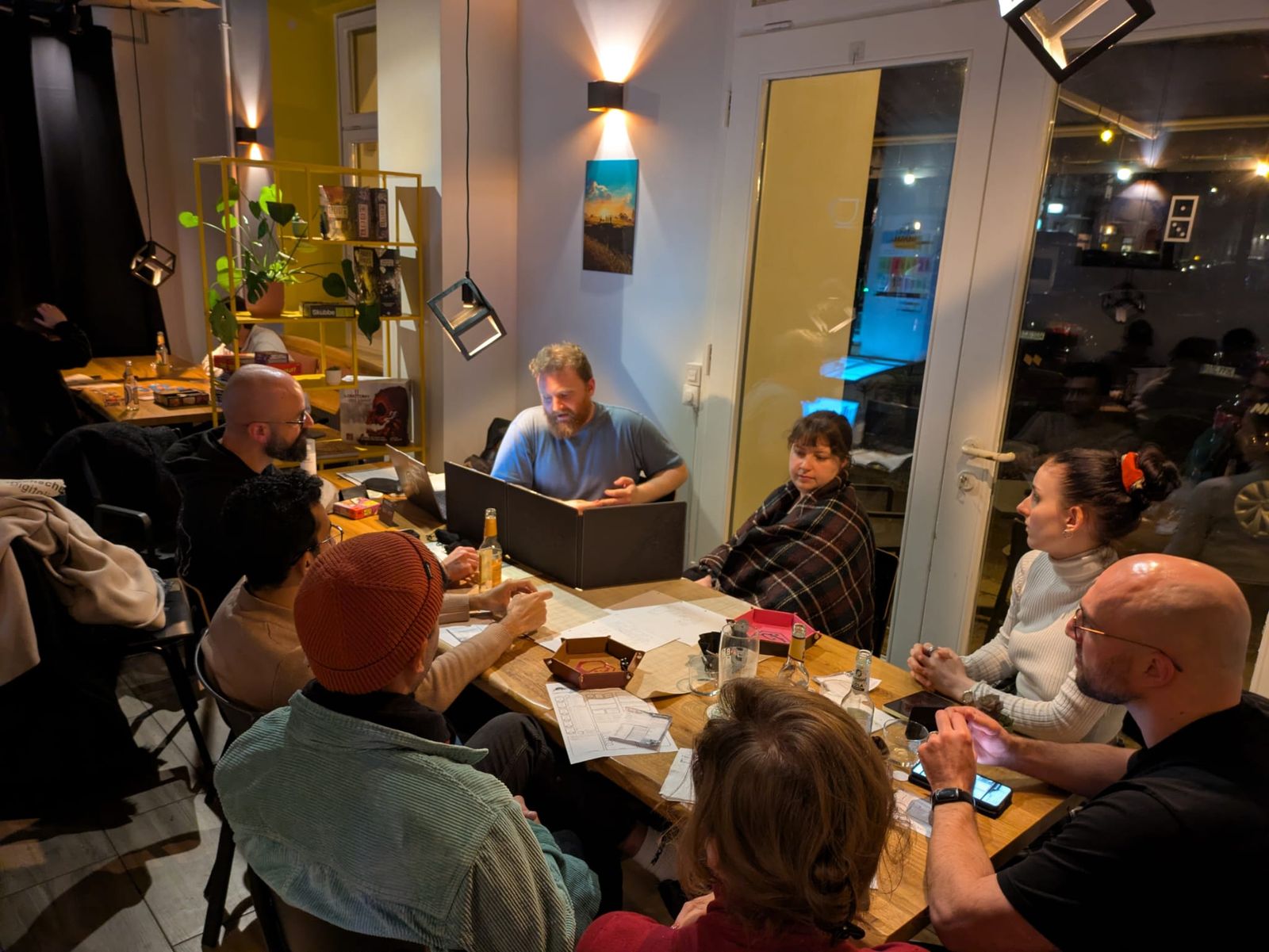 A long table at night, the game master gesturing from behind a screen while eight players listen.