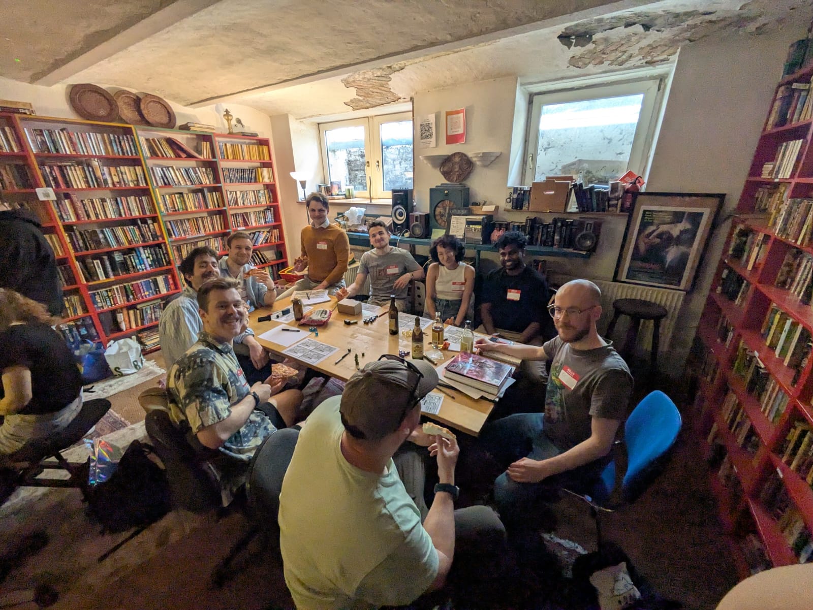 A long table of players under red bookshelves and a low ceiling, bottles and character sheets spread between them.